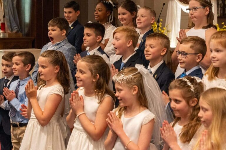 Children in formal attire gathered for a First Communion ceremony, smiling and clapping.