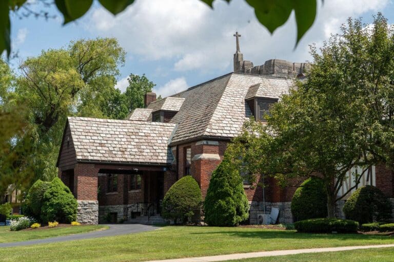 Brick church with stone roof surrounded by trees and greenery under a blue sky.