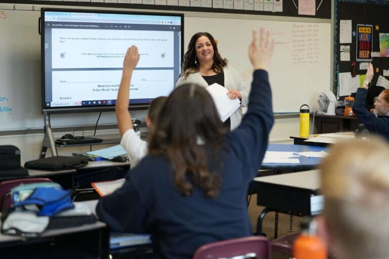 Teacher leading a math lesson with interactive questions in a classroom setting; students eagerly participate.